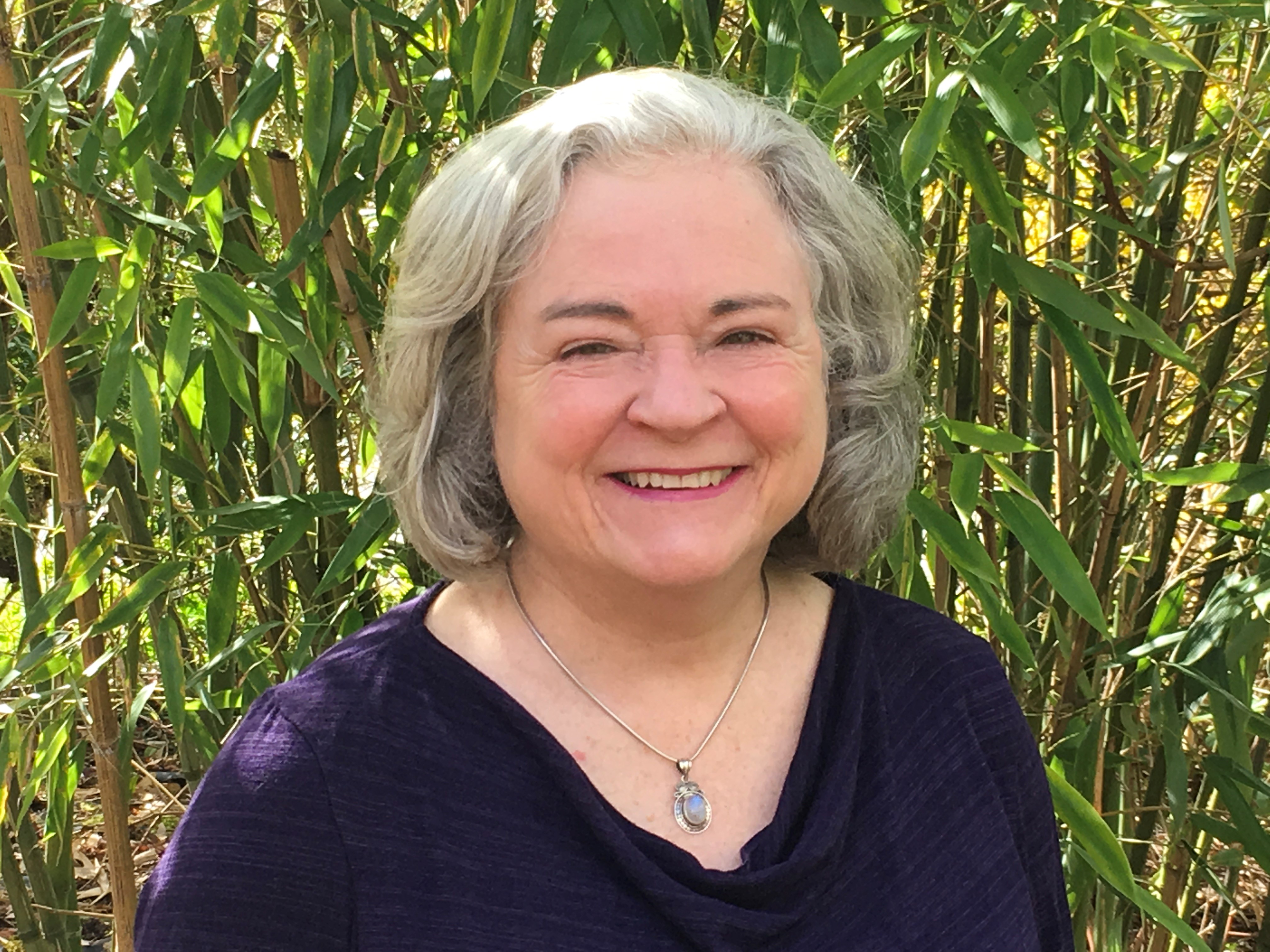 Image: Trish Borden, an aged white woman with gray hair and a purple blouse smiles.