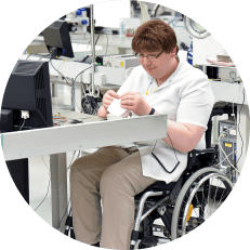 Image: Circular photo of a man who uses a wheelchair sitting at a tech assembly station, assembling something with his hands.