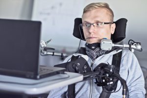 Image: Student who uses a wheelchair and associated equipment looking at a computer, sitting at a desk.