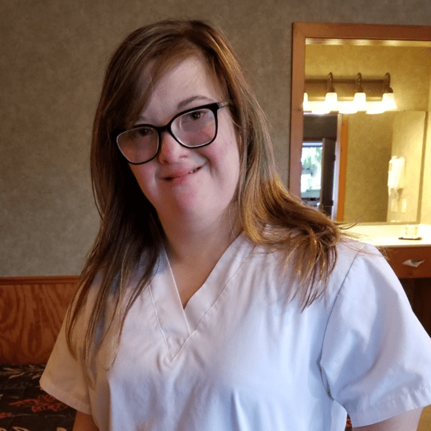 Image: Young woman with ginger hair wearing black framed glasses and white scrub top smiles.
