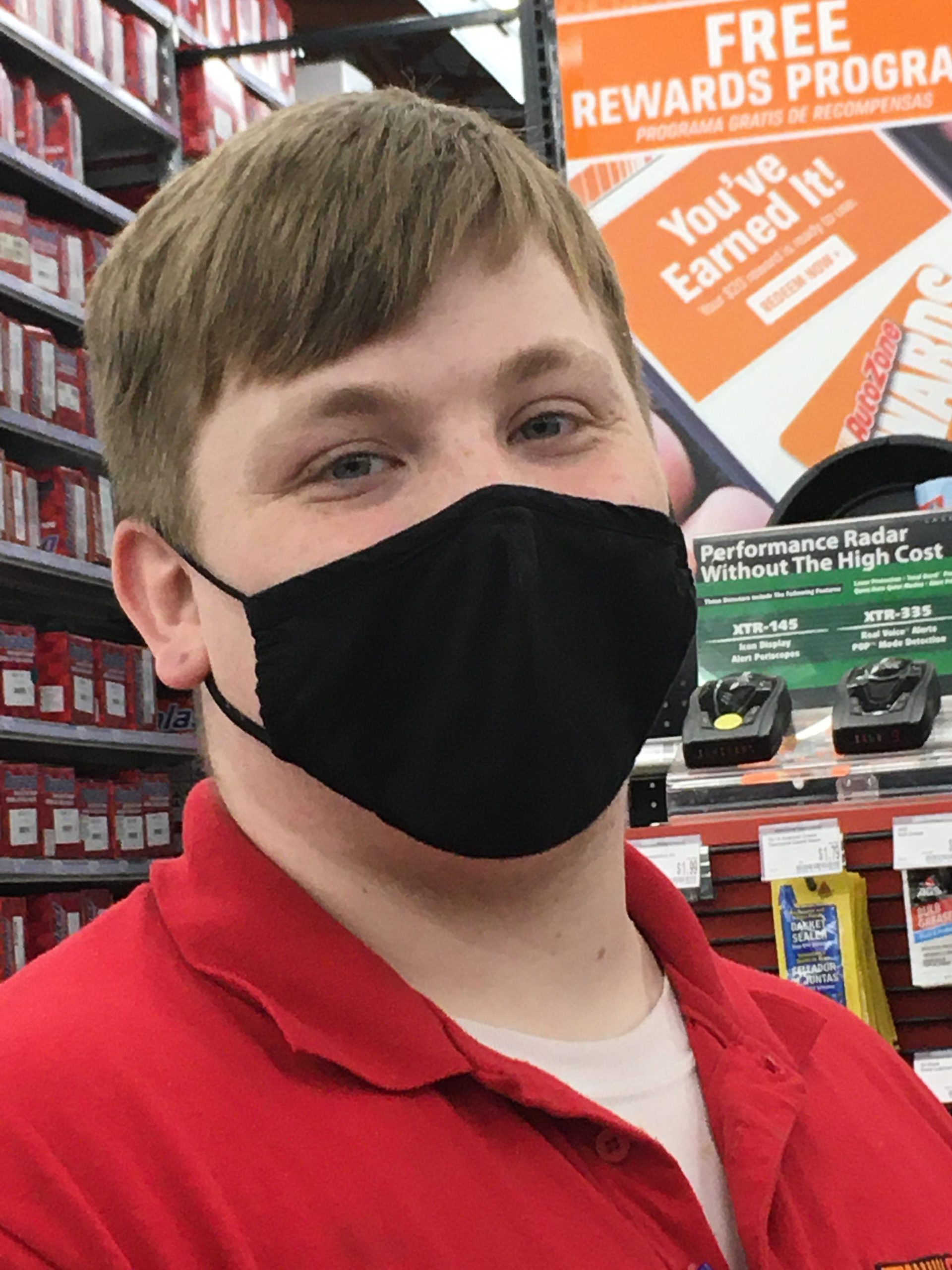 Image: Man with short brown hair, black mask, and red shirt looks into the camera. Photo is from chest up.