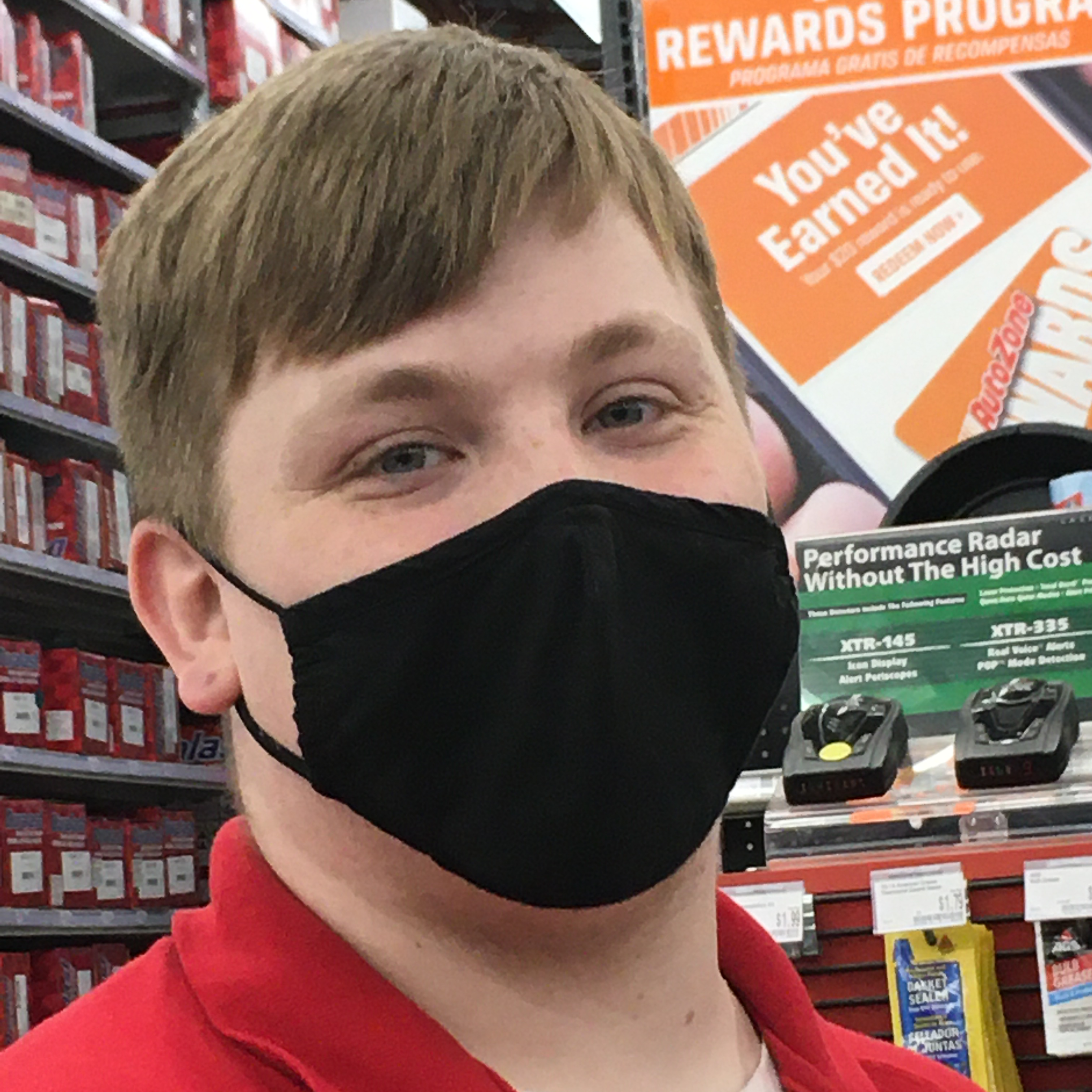 Image: Man with short brown hair, black mask, and red shirt looks into the camera. Photo is from chest up.