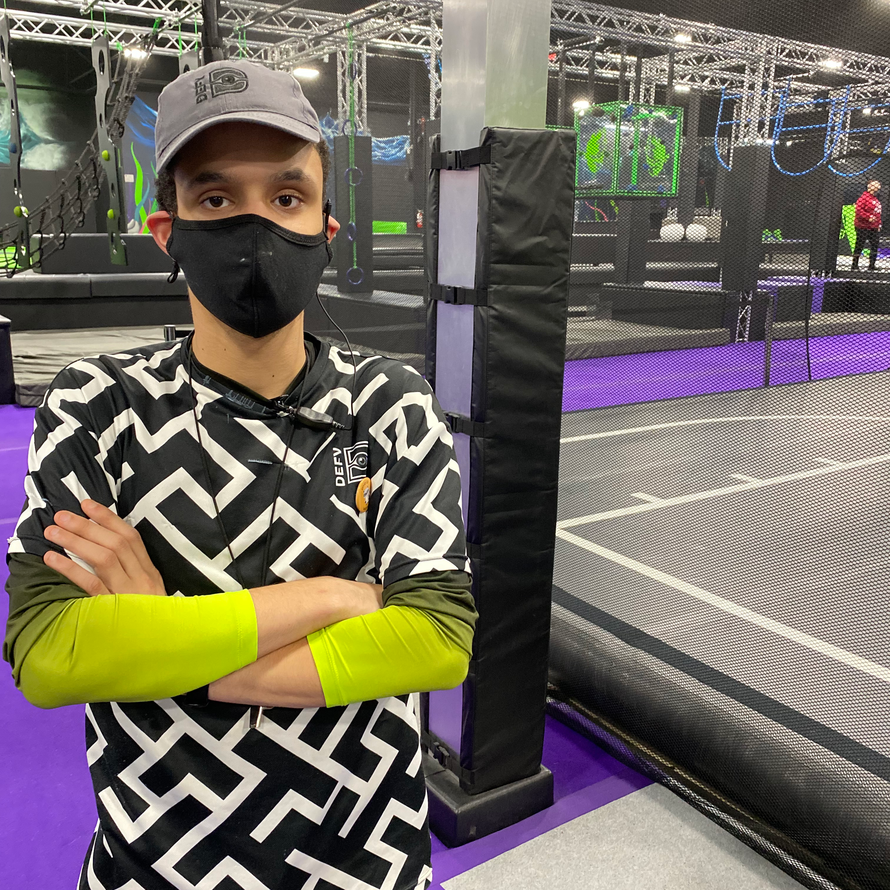 Image: Man with black mask and hat folding his arms with his work uniform on. Black and white short sleeve shirt in a maze pattern with a lime green long sleeved shirt underneath.