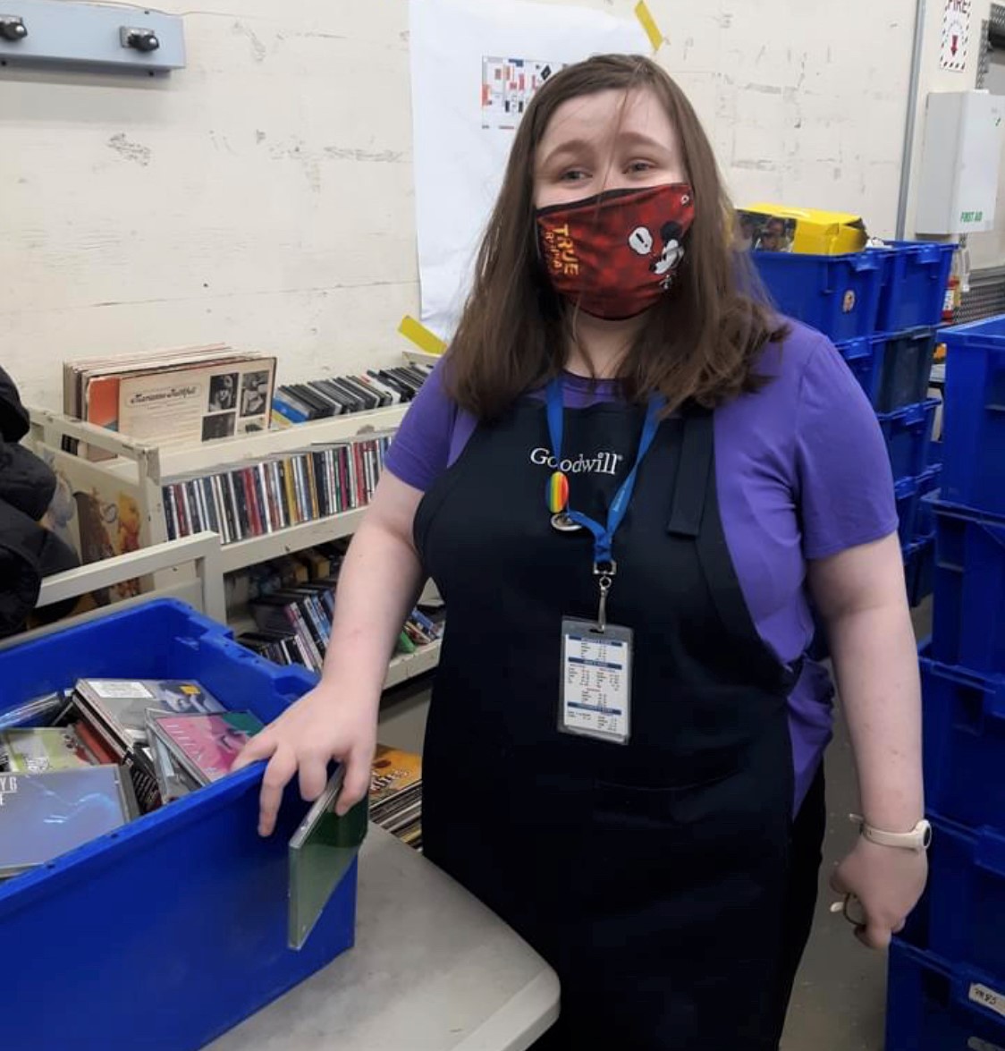 Image: woman with brown hair, bergundy mask, purple shirt, and black Goodwill apron stands in front of shelves of DVD's.