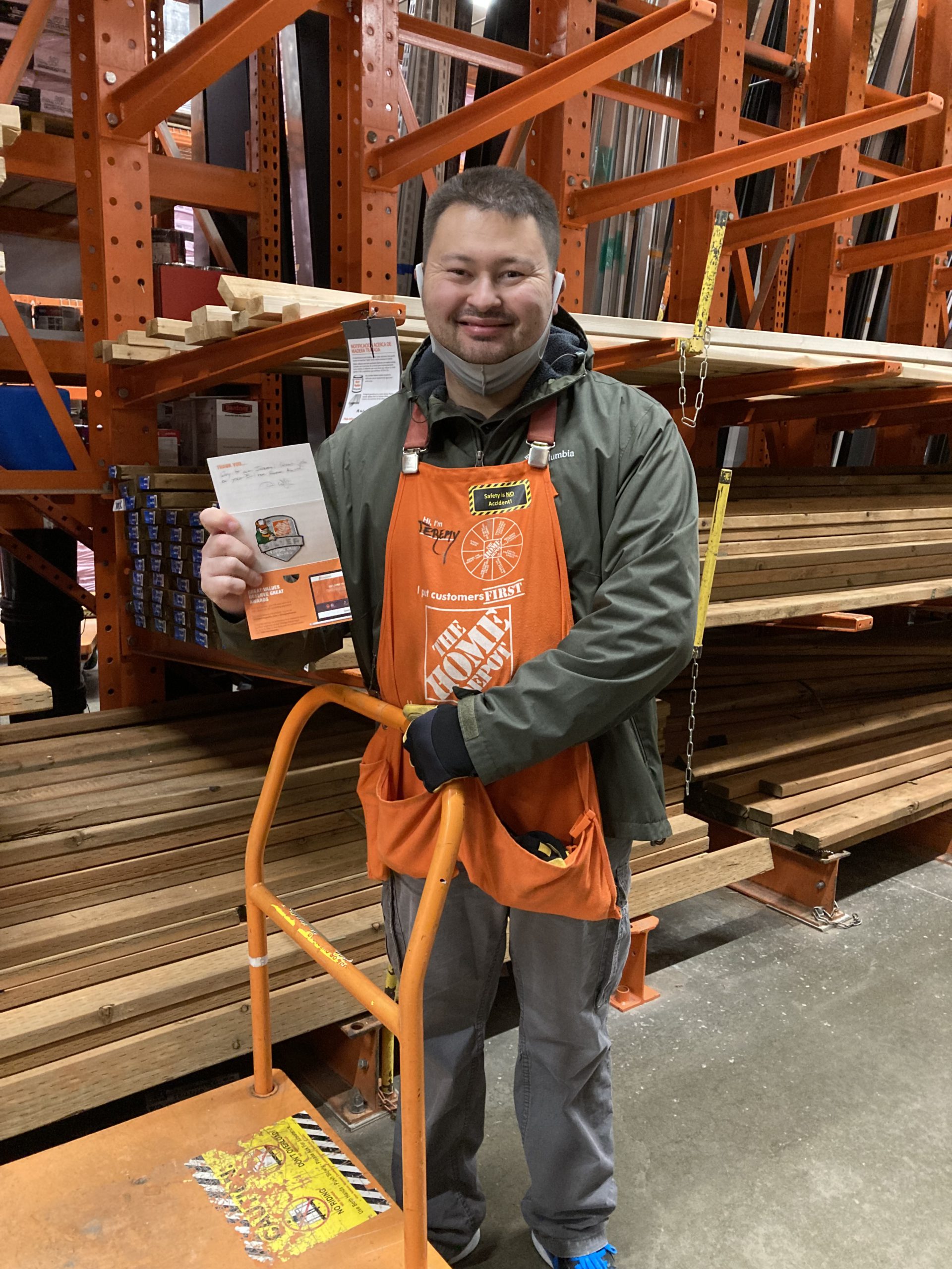 Image: Man with short brown hair wearing a Home Depot apron smiles, holding a card in one hand, his other hand on the handle of a Home Depot cart. He is standing in front of lumber.
