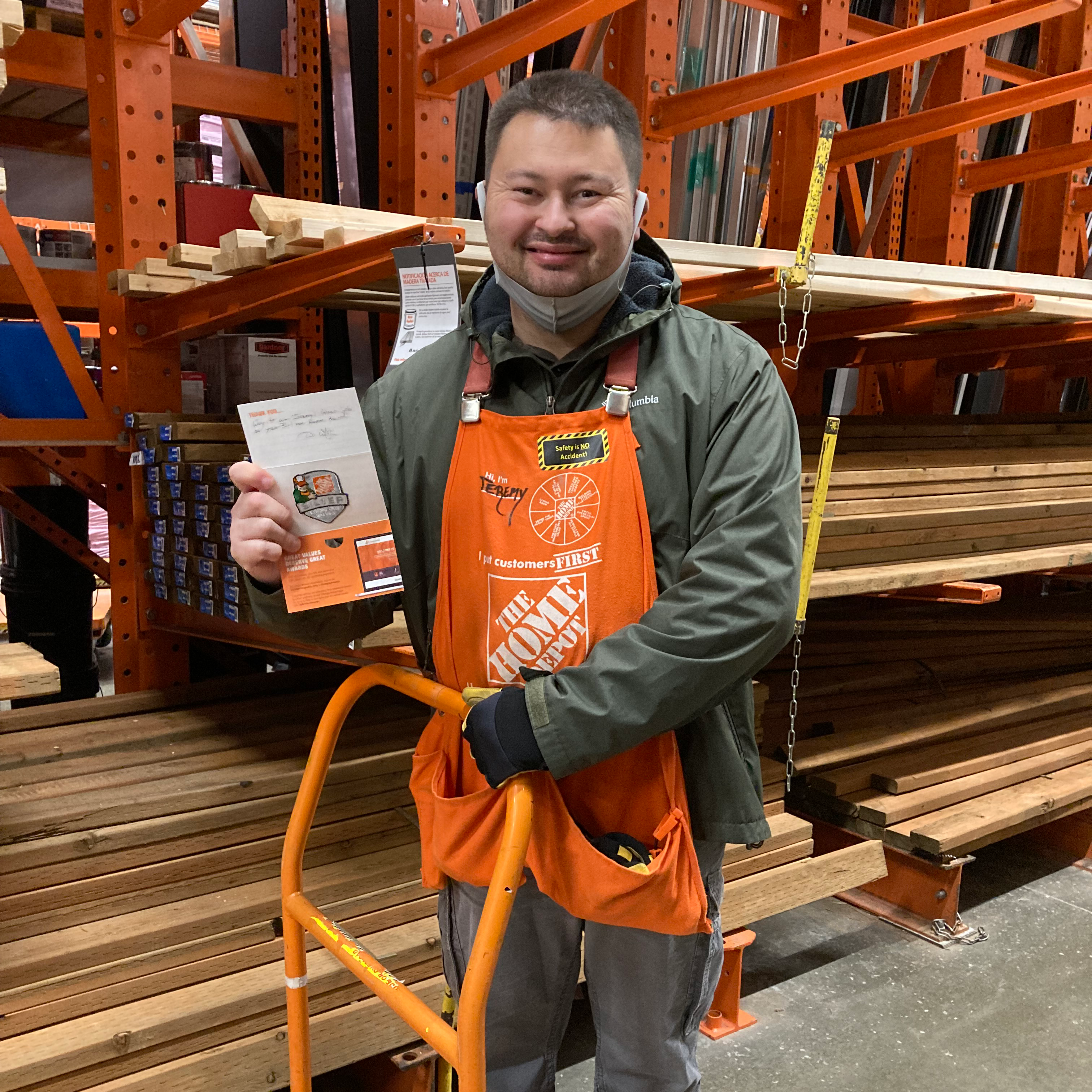 Image: Man with short brown hair wearing a Home Depot apron smiles, holding a card in one hand, his other hand on the handle of a Home Depot cart. He is standing in front of lumber.
