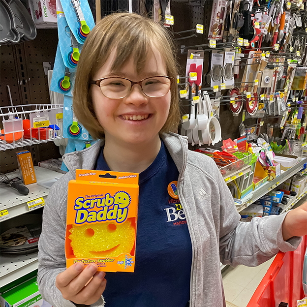 Image: Naomi stands in the cleaning aisle at Henery Hardware, smiling and holding a "Scrub Daddy", a smiley face scrubber. She has brown hair cut above her shoulders, glasses, and is wearing a dark blue t-shirt under a light gray hoodie.