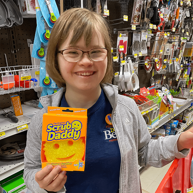 Image: Naomi stands in the cleaning aisle at Henery Hardware, smiling and holding a "Scrub Daddy", a smiley face scrubber. She has brown hair cut above her shoulders, glasses, and is wearing a dark blue t-shirt under a light gray hoodie.