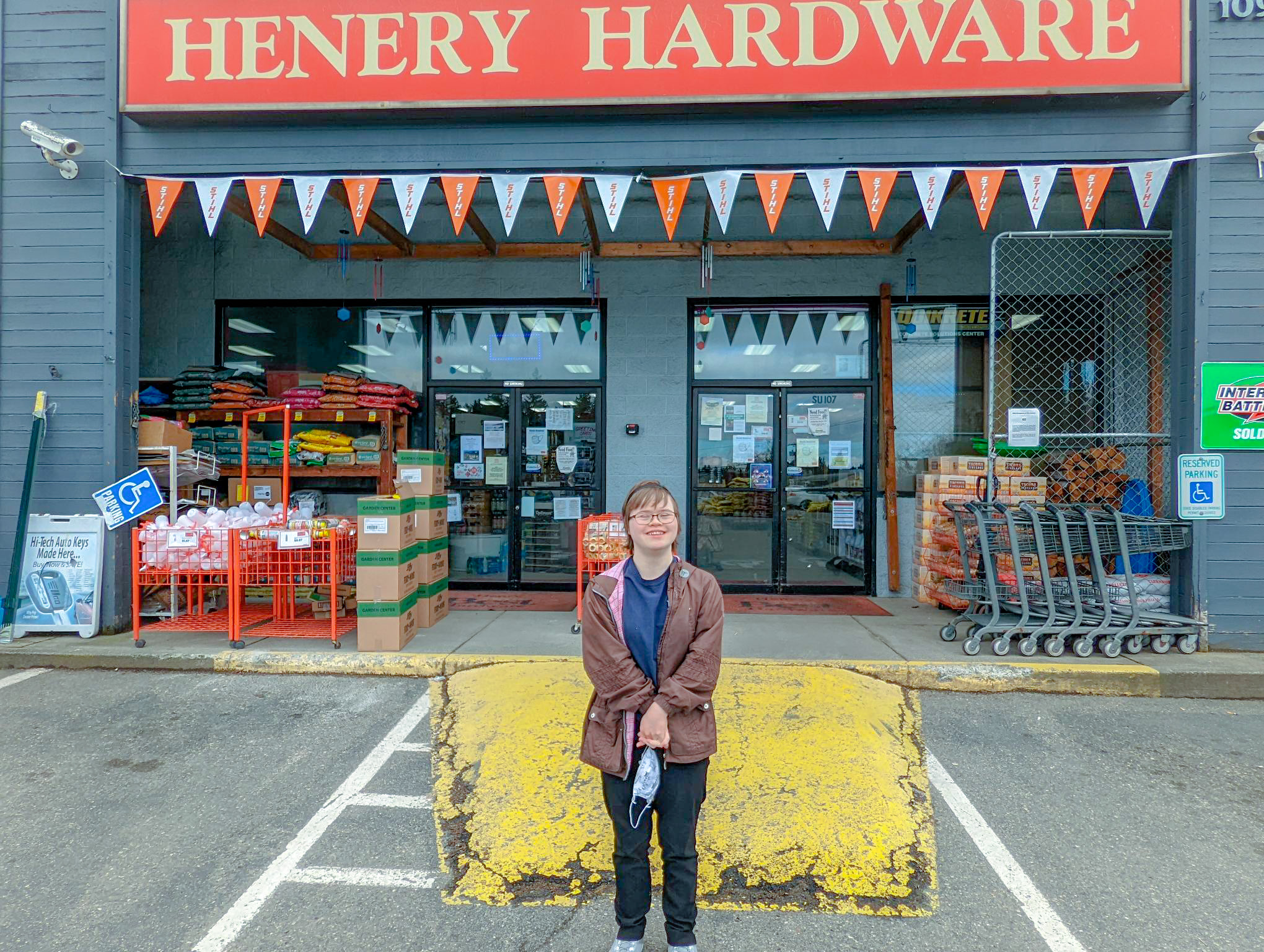Image: Woman with brown coat and mask in her hand smiles in front of Henery Hardware store.