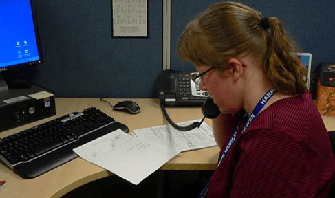 Image: Woman with brown hair and bergundy shirt sits at a computer and holds a telephone receiver to her ear.