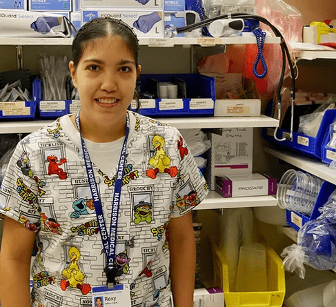 Image: Woman with balck hair, Sesame Street Scrubs, and name tag stands in front of a shelf of medical supplies.