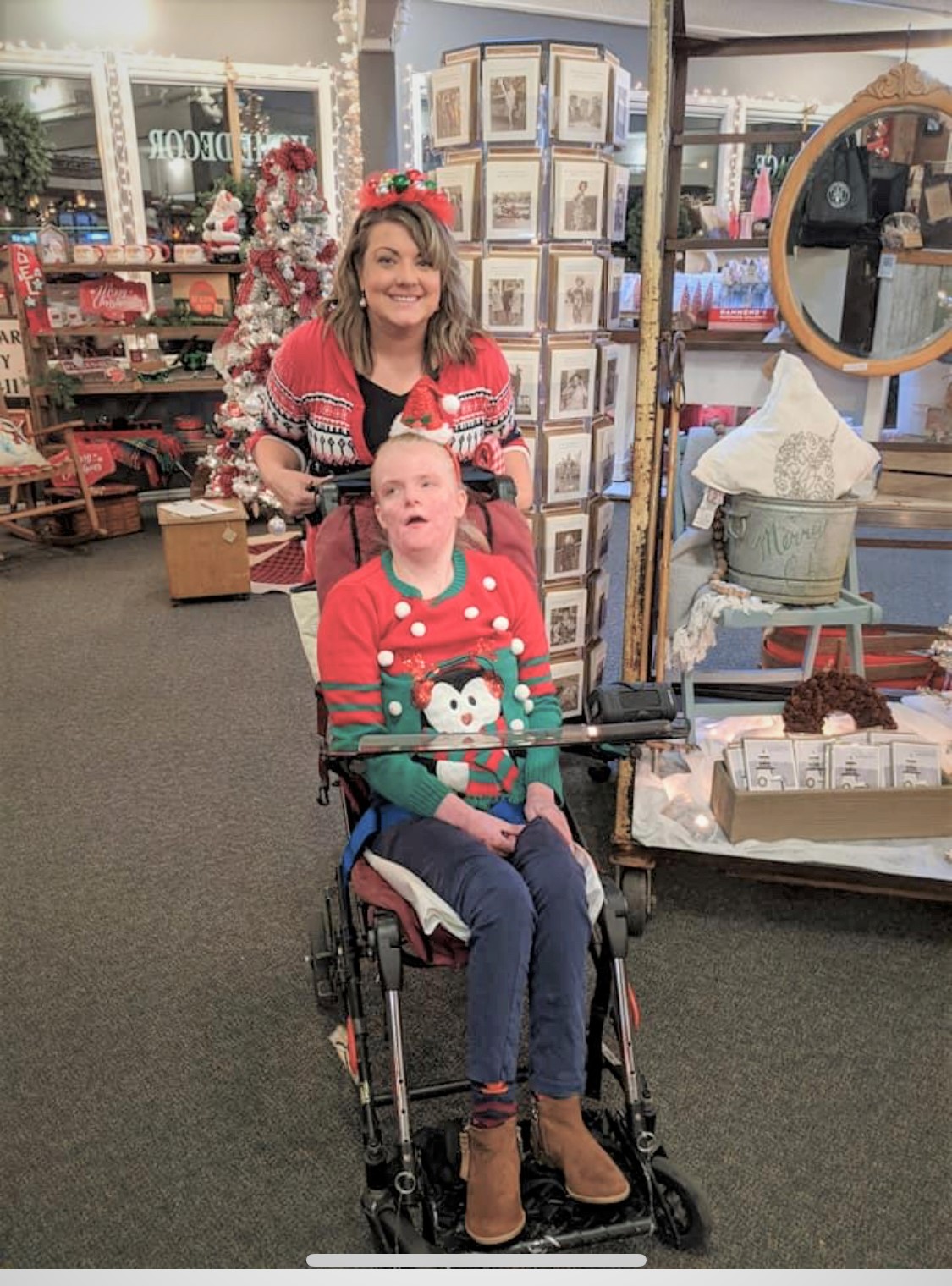 Image: Woman sitting in a wheelchair wearing a Christmas sweater with a penguin on it. Her Job Coach stands behind her, also dressed in a red christmas sweater. They apeear to be in a christmas store with cards and decorations behind them.