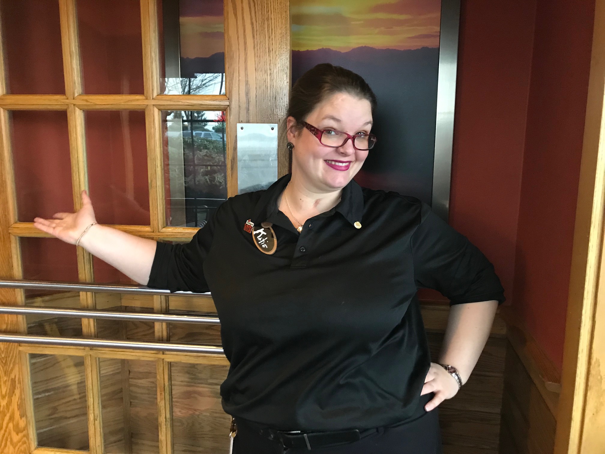 Image: Woman holding a door open and smiling with her right arm showing the way into the door. She wears a black Applebees uniform and name tag.