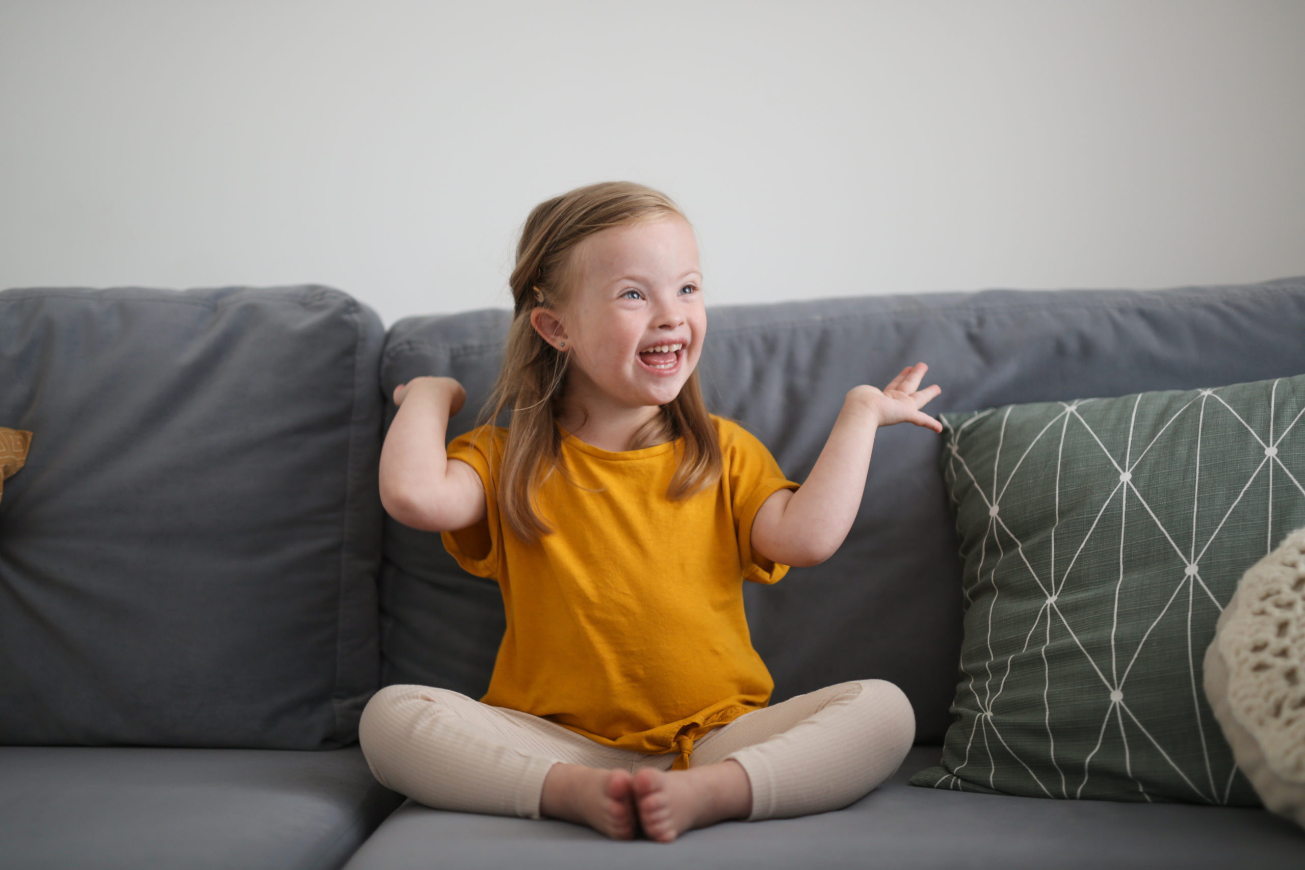 Image: 3 year old girl with Downs Syndrome, blonde hair, and yellow shirt sitting on a gray couch and smiling with hands in the air.