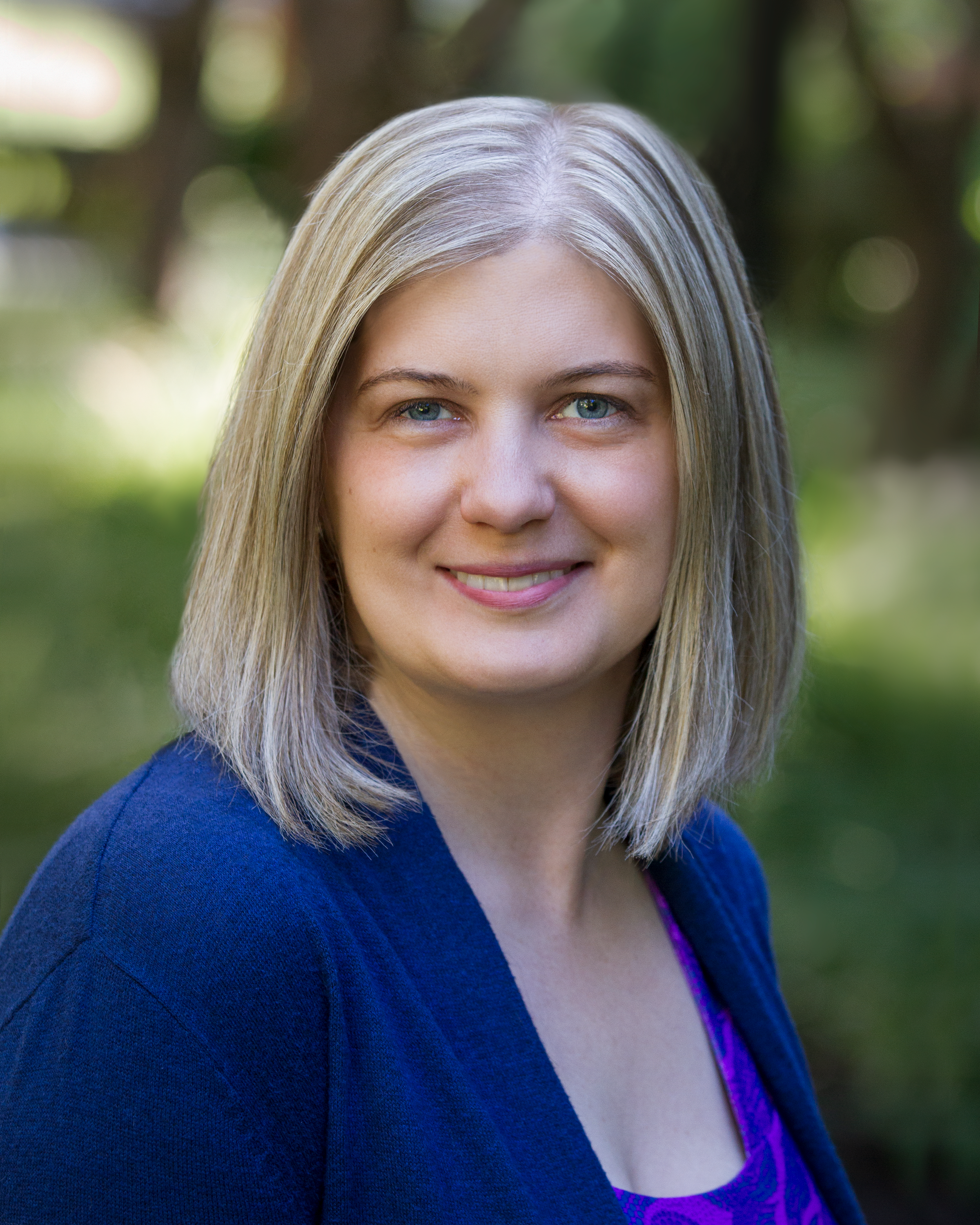 Angela Image: Photo of Angela Dirk, a white woman with shoulder-length blonde hair and blue eyes, wearing a red and blue patterned shirt with a blue cardigan.