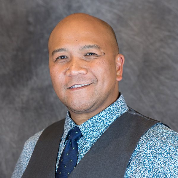 Image: Wally Tablit, a bald Filipino man wearing a blue patterned shirt and dark blue tie and vest smiles.