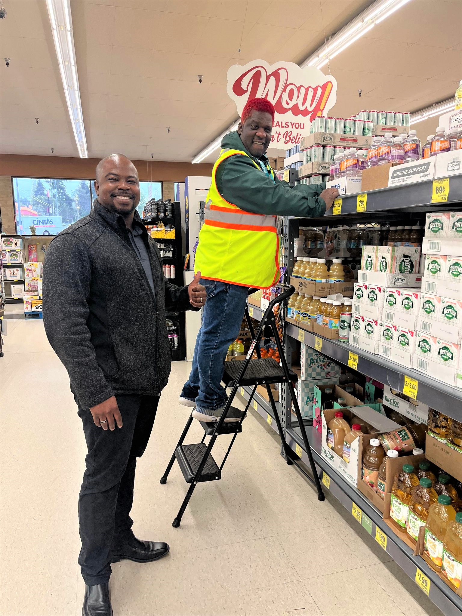 Image: Two men in a Grocery Store aisle. One stands on a step ladder wearing a high-visibility vest and stocks shelves. The other man stands behind him smiling at the camera and giving a thumbs-up.
