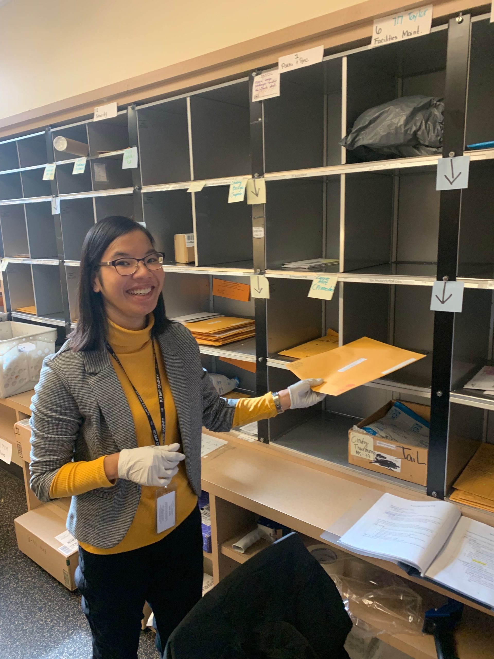 Image: Woman with black hair passed her shoulders, glasses, a yellow shirt, gray blazer, latex gloves, and a badge hanging around her neck on a lanyard, puts mail into a wall organizer.