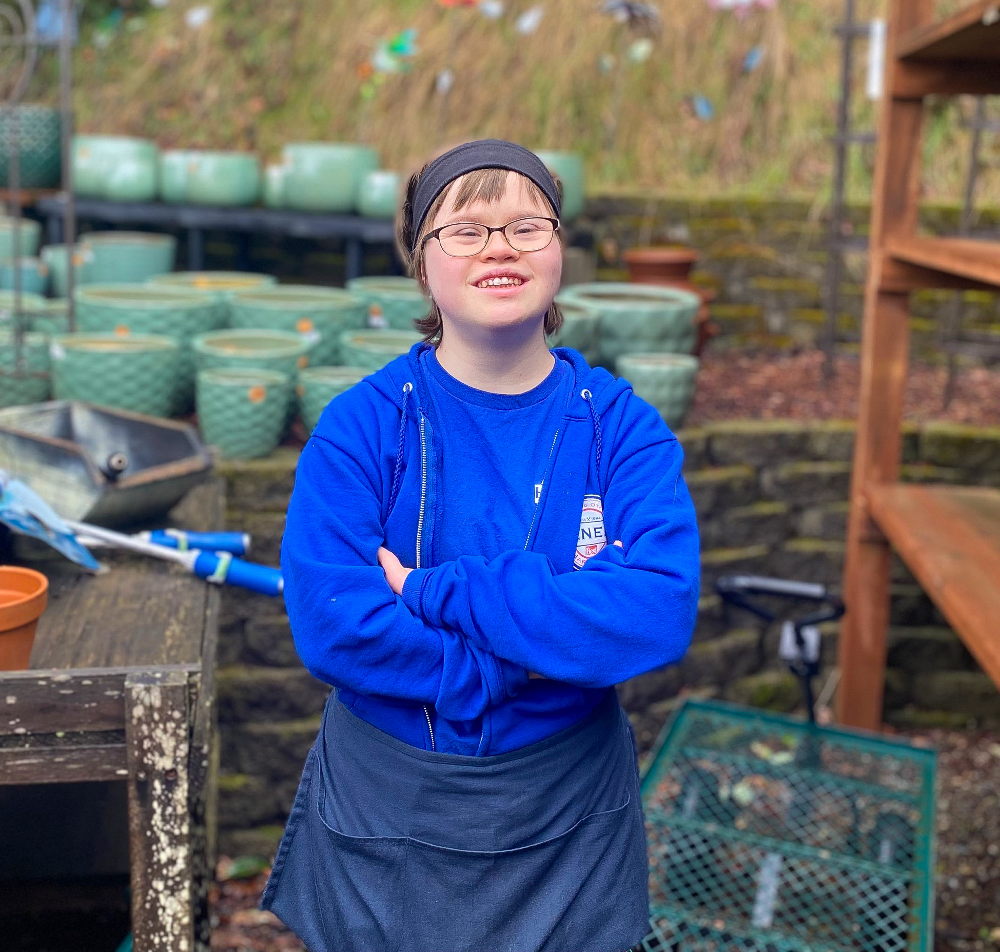 Image: Naomi stands outside in front of a table of empty blue ceramic flower pots with her arms crossed and smiling, wearing a royal blue hoodie & t-shirt, black-rimmed glasses, and shop apron.