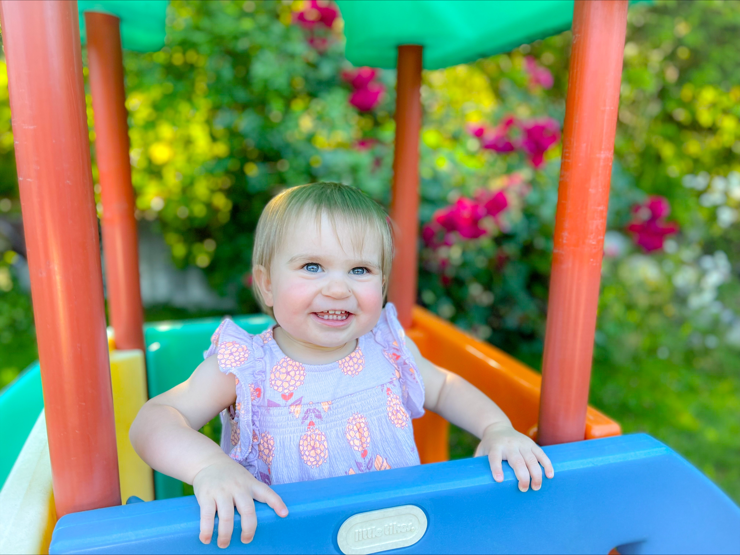 Image: Toddler with blonde hair and blue eyes smiles from inside a multi-colored outdoor playset.