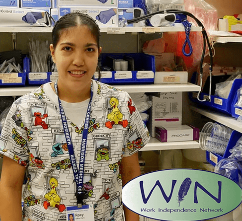 Image: Woman with black hair pulled back, wears colorful scrubs and smiles, standing in front of a shelf of medical supplies.