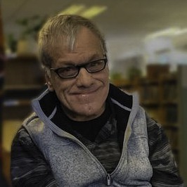 Image: Jeff Grace, a white man with dark-rimmed glasses and gray hair wearing a light blue vest over a dark shirt, smiles.