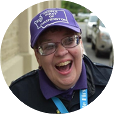 Image: Resa Hayes, a white woman with a dark jacket, glasses, and a purple baseball cap smiles.