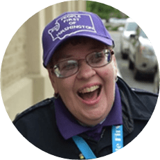 Image: Resa Hayes, a white woman with a dark jacket, glasses, and a purple baseball cap smiles.