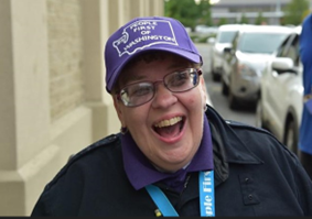Image: Resa Hayes, a white woman with a dark jacket, glasses, and a purple baseball cap smiles.