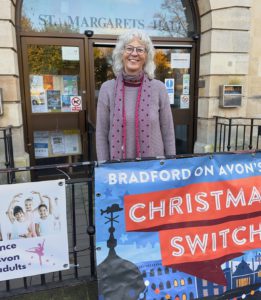 Anne is standing in front of the community center in Bradford on Avon where she lives. There are lots of posters of community and business activities in the picture. She is wearing a gray sweater and polka dot scarf, has shoulder length hair and is smiling at the camera.