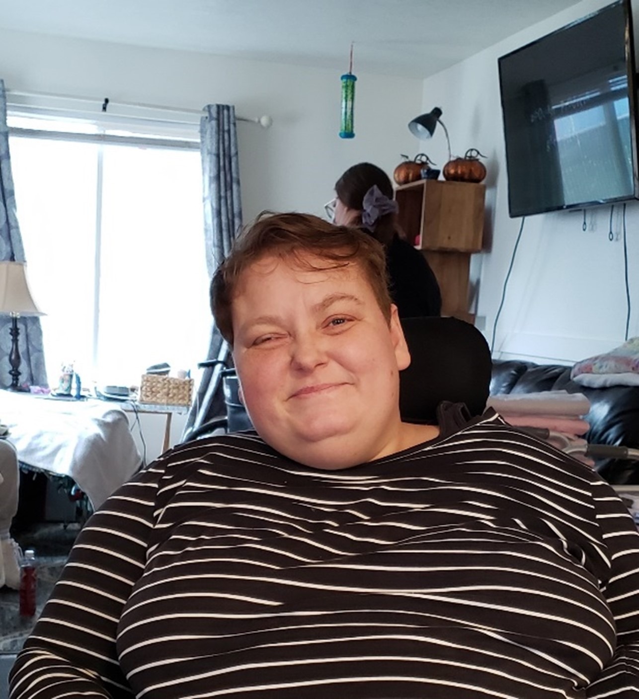 Description: Annette is sitting in front of a window and television. She is wearing a white and black striped long sleeve shirt, has short brown hair, and is smiling at the camera.