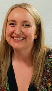 Kayla is sitting in front of a white blank wall. She is wearing a black shirt that has a red, yellow, and green floral pattern and is wearing gold hoop earrings. She has long blonde hair and is smiling at the camera.