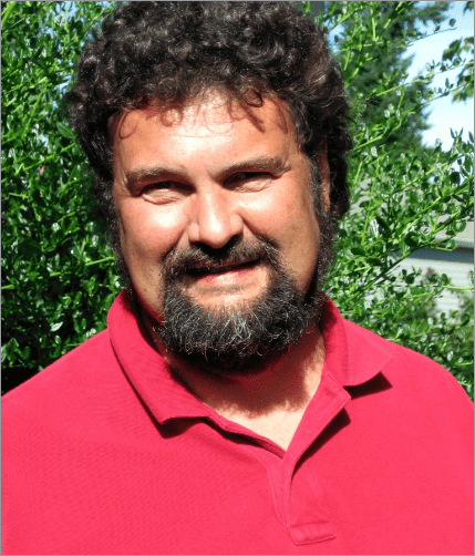Image: Chuck Goodwin, a white man with dark brown curly hair and beard, wearing a red Polo shirt, smiling and slightly squinting in the sun.